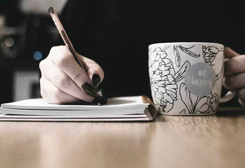 A person writes in a notebook and drinks something from a cup